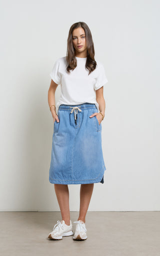 Woman wearing a Mid Blue light weight Denim Skirt with elasticated wait and drawstring standing against a white background