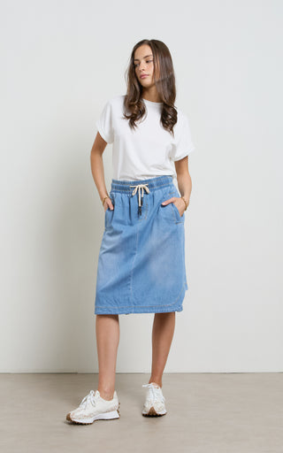 Woman wearing a Mid Blue light weight Denim Skirt with elasticated wait and drawstring standing against a white background