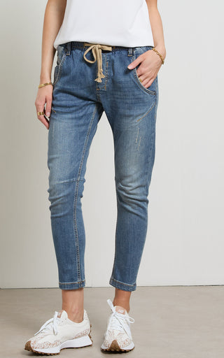 Woman wearing a Mid Blue Denim jogger jeans with elasticated wait and drawstring standing against a white background
