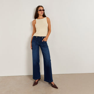 Woman wearing a beige sleeveless top and dark blue jeans against a plain background